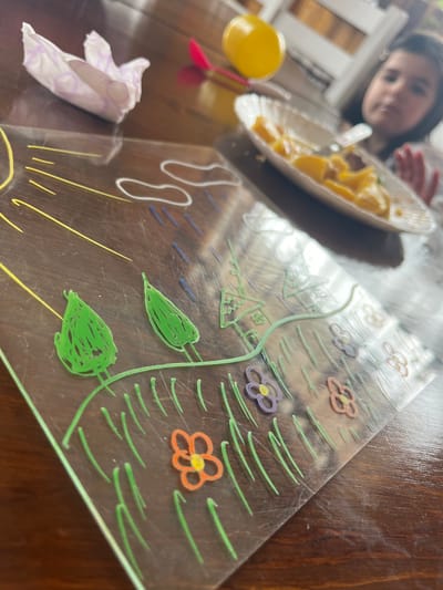 The image shows a table setting with a child and a plate of food partially visible in the background. In the foreground, a clear placemat with a colorful drawn design of flowers, leaves, and sun rays rests on the table. A crumpled tissue sits next to the placemat, and a yellow toy is also present. The table surface appears to be made of wood. There is no information related to Breznik, Bulgaria.