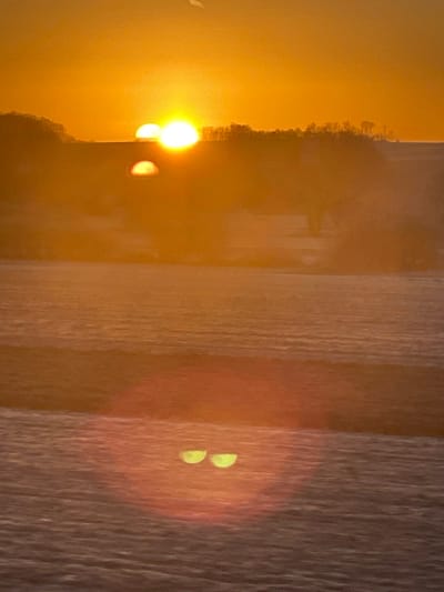 An abstract, warm-toned image features a hazy scene with vertical, textured lines dominating the right side. A bright yellow sun or light source is partially visible on the left, casting a vibrant glow and lens flare that contributes to the overall blurry effect. Two small, bright green, crescent shapes are visible inside the reddish lens flare. The scene is soft, lacking crisp edges, and has a dreamlike quality.