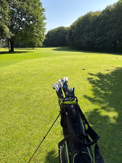 A black golf bag with a lime green trim sits on the 15th green of the Lancaster Golf Club, located at 54.01 latitude and -2.829 longitude. Four golf clubs are resting in the bag. The bag is facing a long green fairway with trees bordering the course on the right. The sun is shining and there are no people visible. The ground is covered in green grass and a long shadow stretches from the bag toward the fairway.