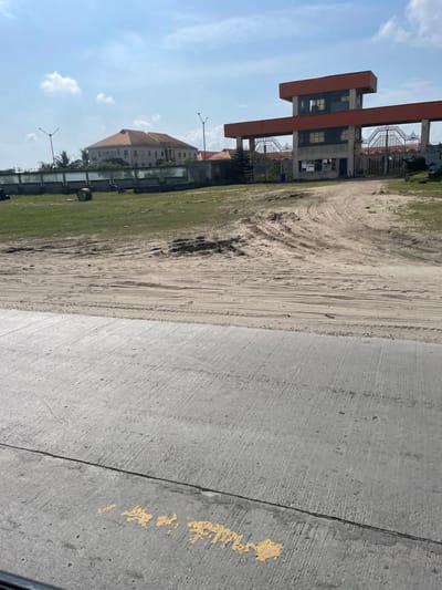 The image depicts an outdoor scene in Igando Oloja, Nigeria, under a bright, partly cloudy sky. In the immediate foreground, a grey concrete surface, possibly a road or paved area, exhibits faint yellow paint markings and a dark, indistinct object at the bottom edge. Beyond this, a wide expanse of disturbed sandy terrain shows numerous vehicle tire tracks and patches of green grass. Lying on the grassy section to the left are several large, dark, circular objects, possibly industrial tires or spools.

In the mid-ground, a prominent multi-level gateway structure dominates the right side. It features a concrete base, an elevated control or observation room with an orange facade, and ornate metal gates that appear closed. A blue waste receptacle is visible near the base of the gate on the left side. Behind this gateway, a building with a reddish-brown tiled roof is partially visible. Further left in the background, another large, multi-story building with a light-colored facade and a matching reddish-brown tiled roof stands behind a low fence or wall. Two streetlights with multiple fixture heads are positioned in the background. No individuals are present in the scene, and no overt actions or interactions are depicted.