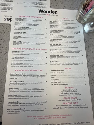 The image shows a menu from Wonder., a restaurant offering "Cool Food Cool Drinks". The menu is divided into several sections including Breakfast Sandwiches, Lunch, Grande Breakfast Burritos, Breakfast Favorites, Sides, Drinks, Cold Beer, and Mimosa Bar. Breakfast sandwiches include Bacon, Egg & Cheese, Sausage, Egg & Cheese, and Mushrooms, Egg & Cheese. Lunch includes options like Spicy Fried Chicken Sandwich, California Club Sandwich, The Standard Cheeseburger, The Panorama Sandwich, and The Topanga Sandwich. Grande Breakfast Burritos offer House Made Chorizo and Smoky Black Beans. Breakfast Favorites include Desert Vegetarian Bowl, Wonder Big Breakfast, Avocado Toast, French Toast, Loaded Hash Browns, and Holli's Homemade Granola. Sides include Two Hash Brown Patties, Seasoned French Fries, Avocado, Bacon, Sausage Patty, Sourdough Toast, and Two Fried or Scrambled Eggs. Beverages offered are Coke, Diet Coke, Sprite, Sparkling Water or Still Water, Iced Black Tea, Arnold Palmer, and Orange Juice, Strawberry Lemonade. Cold beer options are Stella Artois, Peroni, Heineken, and Modelo. The Mimosa Bar offers Orange, Strawberry, Peach or Pineapple Mimosa. The menu states that items are prepared as described, with no modifications allowed, and that a $5 charge will be added to all shared plates.