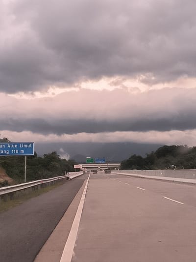 The image presents a view from a moving vehicle on a multi-lane highway in Seulimeum, Indonesia. The road, featuring a solid white line on the left shoulder and dashed white lane markings, stretches into the distance. A metal guardrail runs along the left side, while a concrete barrier borders the right, partially obscuring a larger vehicle.

The sky is heavily overcast with dark, dense clouds, giving a muted and slightly reddish-grey hue, particularly where light filters through the upper cloud layers. This suggests either a very cloudy day, or possibly early morning or late afternoon, with potential for rain. Lush green trees and foliage line the highway, and dark, hazy mountains or hills are visible in the background, partially obscured by the low-hanging clouds and a faint mist.

In the distance, a few cars are seen driving on the highway. A blue highway sign on the left provides location information, partially reading "an Alue Limut" and "jang 110 m," which likely refers to "Simpang Alue Limut" (Alue Limut Intersection) at a distance of 110 meters. The overall scene conveys a feeling of travel through a verdant, mountainous region under dramatic, cloud-filled skies. No people are explicitly visible.