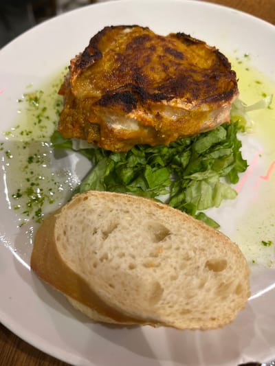 A white plate contains a single serving of food, likely a Basque tapas dish, consisting of a fried, round piece of food on a bed of lettuce, accompanied by a slice of bread. The plate contains green, what appears to be parsley, oil and water droplets, and the plate is slightly tilted upwards. The coordinates 43.324, -1.986 indicate the location of the city of Donostia, Spain. The scene is a close-up of the dish on a table. There is no indication of who might be dining on the food.