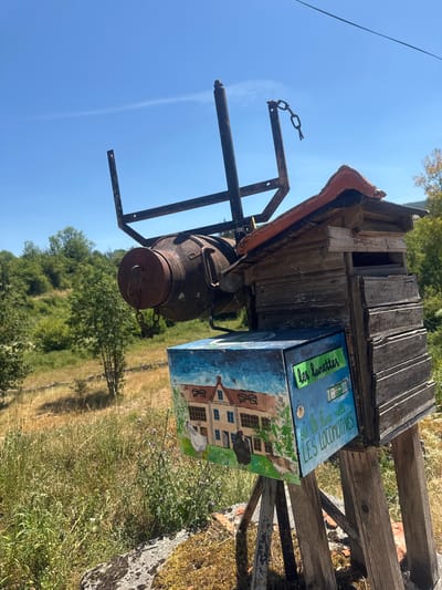 This is an outdoor, eye-level, full shot of a rustic, handmade mailbox situated on a hillside in Coteaux du Lizon, France. The mailbox is a multi-component structure. The main body appears to be constructed from weathered, horizontal wooden planks, topped with a section of red tile roofing. Attached to the top is a rusty metal frame with a chain hanging from it, and a large, rusty, cylindrical object resembling a boiler or industrial part suspended by the frame. A smaller, square box, painted with a pastoral scene of a house and chickens, is attached to the front of the wooden structure. The box has text written on it that reads "les lunettes" and "si la lune naît LES LOCOMOTIVES". The background consists of a dry, grassy hillside with scattered trees under a clear blue sky with a faint wispy cloud. The scene is well-lit by natural sunlight. There are no people or animals visible in the frame other than those depicted in the painting on the mailbox.