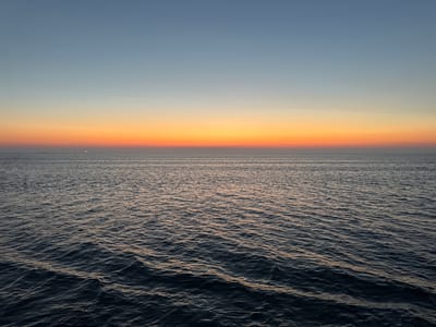 The image depicts a wide expanse of the ocean at dusk. The sky transitions from a pale blue at the top to a vibrant orange and pink band just above the horizon, indicating the setting or rising sun. The sea is a deep, dark blue with visible ripples and waves, reflecting the colors of the sky. In the far distance, on the horizon line, there are a few small specks that appear to be ships. The scene is devoid of any identifiable landmarks or structures that would suggest a specific location within the city of Fehmarn, Germany. The focus is solely on the natural elements of the sky and sea.