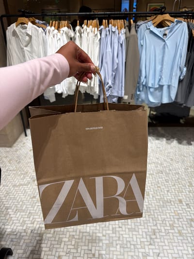 A person holding a Zara shopping bag is standing in front of a clothing rack. The bag is brown paper with the Zara logo printed in white, and it appears to be made of 100% recycled paper. The clothing rack is full of shirts in various colors, including blue, gray, and white. The floor is covered in a white herringbone pattern tile. The scene takes place in a Zara store, likely located in the city of Cerritos, United States of America. The person's arm is visible, wearing a long-sleeved pink shirt. The person's hand is holding the bag's handles.