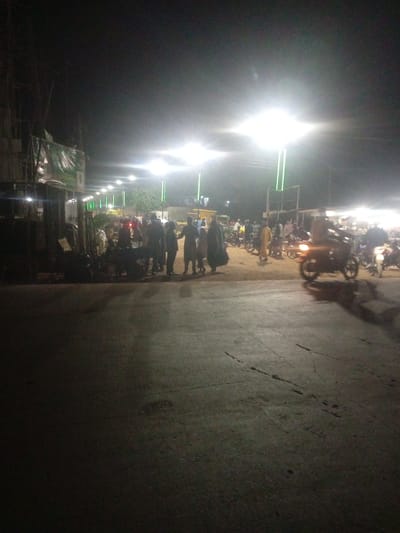 The image depicts a bustling street scene at night in Tundun Wada South, Nigeria. The primary subjects are a crowd of people gathered together, alongside several motorcycles, suggesting a lively evening event or gathering. The setting appears to be an outdoor market or public space illuminated by bright overhead lights, casting a strong glow and creating dramatic shadows.
The people are seen standing and interacting, some appearing to be in conversation. They are dressed in various attire, with some individuals wearing traditional clothing. The motorcycles, parked and in motion, indicate a common mode of transportation and possibly a recreational event, such as a parade or informal gathering.
Notable details include the time of day, clearly night, due to the artificial lighting and dark sky. The bright lights suggest clear weather. The presence of multiple people and motorcycles suggests a community event, possibly a local festival or a busy marketplace operating after dark. The atmosphere seems energetic and social.
No visible text is discernible in the image. The overall impression is of a vibrant community coming together at night.