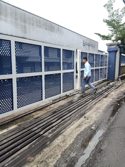A man in a light blue shirt and jeans is walking through an open gate with a blue and gray grid pattern. The gate is attached to a large, gray building. Below the gate is a series of horizontal slats running across the passageway. A section of wet pavement is visible in the foreground, leading to a road. The sky is overcast. Trees are visible in the background behind the gate.