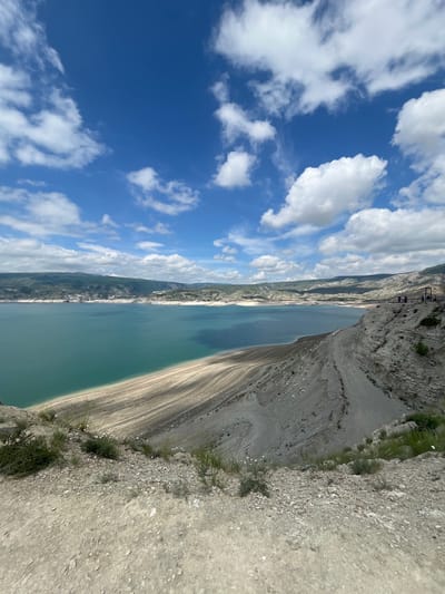 The image depicts a view of the Chirkey Dam reservoir in Chirkey, Russia, photographed from an elevated position along the embankment. The reservoir’s turquoise water stretches horizontally across the frame. To the left, the sky is visible with scattered white clouds against a blue background. On the right side of the image, a rocky embankment slopes down to the water’s edge. In the upper left, a construction or maintenance structure is visible at the edge of the frame. The image shows land erosion, and water level drop.