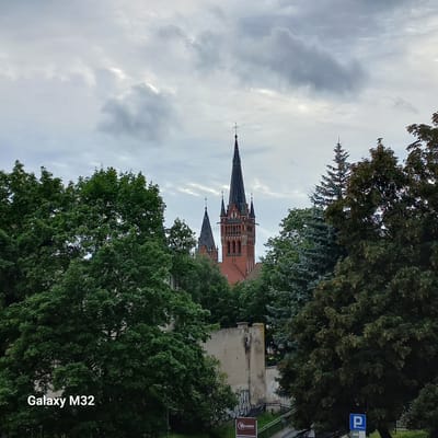 A medium shot captures the spires of a red brick church against a cloudy sky, framed by dense green trees. The church features a tall, dark spire topped with a cross, and a shorter, similarly shaped spire to its left. A clock face is visible on the main spire. In the foreground, the lower branches and leaves of lush trees obscure the base of the church and a light-colored building with graffiti on its lower section. A sign with a blue "P" for parking is visible near the bottom right corner. The "Galaxy M32" watermark is present in the bottom left corner. This scene is likely within the city of Olsztyn, Poland, given the camera watermark, though specific landmarks are not definitively identifiable from the image alone.
