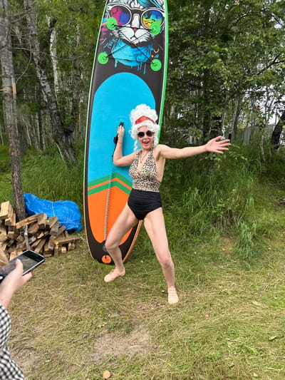 A person dressed as an elderly woman, complete with a white wig, red headband, sunglasses, and a leopard-print swimsuit with high-waisted black bottoms, stands next to a stand-up paddleboard. The paddleboard is decorated with a cartoon cat wearing sunglasses and has bright blue and orange sections. The person is in a playful pose, with one arm outstretched and the other holding onto the paddleboard. They are wearing light pink shoes. To the left of the paddleboard, there is a pile of chopped firewood and a blue tarp. In the foreground, a person's hand is visible holding a smartphone, as if taking a photo. The scene is set in an outdoor environment with trees and grass, suggesting a rural or park-like setting. The location is identified as Tolbazikha, Russia.