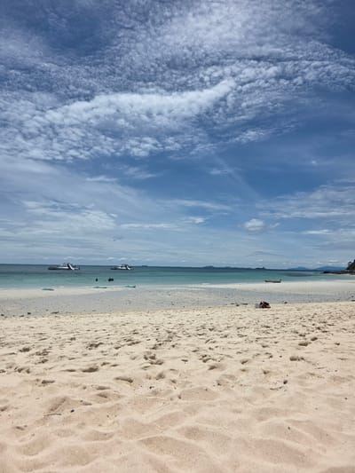 The image depicts a beach scene in Bang Sare, Thailand. The perspective is from the sandy shore, looking out towards the ocean and sky. The sand is light-colored and shows imprints of footprints, suggesting recent activity. In the foreground on the right side of the image, there is a bright orange life vest and a black bag, possibly belonging to a beachgoer.
Further out, the turquoise ocean stretches towards the horizon, where a line of distant land with hills is visible. Several boats are dotted across the water, with two larger boats appearing to be closer to the shore. The sky above is a vibrant blue, filled with a variety of white clouds. Some clouds are wispy and spread out, while others are more textured and puffy, creating interesting patterns.
The scene suggests a clear and pleasant day, likely during daylight hours given the brightness and cloud formations. The presence of the beach, ocean, and boats points to a coastal location, specifically identified as Bang Sare, Thailand. There are no visible signs of text in the image. The overall mood is serene and captures the essence of a tropical beach environment.