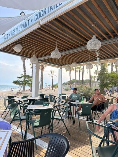 This is a medium shot photo of a beachside restaurant in Cambrils, Spain. The restaurant is covered by a white awning and has a wooden slatted roof. There are several tables and chairs, with two men sitting at one of them. The man on the left is wearing a green t-shirt and a black baseball cap, while the man on the right is shirtless. There is a bottle of beer and a glass on their table. In the background, there is a sandy beach with palm trees and the ocean. Several people are visible on the beach, some walking, some sitting. The sun is setting, casting a warm glow over the scene. The restaurant has a sign that reads "BAR RESTAURANT COCKTAILS".