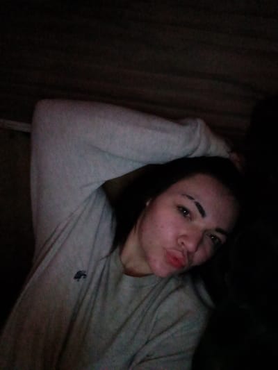 An individual, appearing female, is depicted indoors in a dimly lit environment. The subject is in a reclined position, facing the camera with a direct gaze and pursed lips. She wears a light gray long-sleeved crew-neck shirt with a dark, embroidered emblem on the left chest. Her right arm is bent, with the hand positioned near the upper head/neck region. Her left arm extends towards the lower-left portion of the frame. The subject has dark hair, prominent dark eyebrows, and visible blemishes on her face. The background is dark and indistinct, showing textured surfaces consistent with an interior setting, possibly wood or fabric. No visual information within the image identifies the specific location as Ozark, United States.
