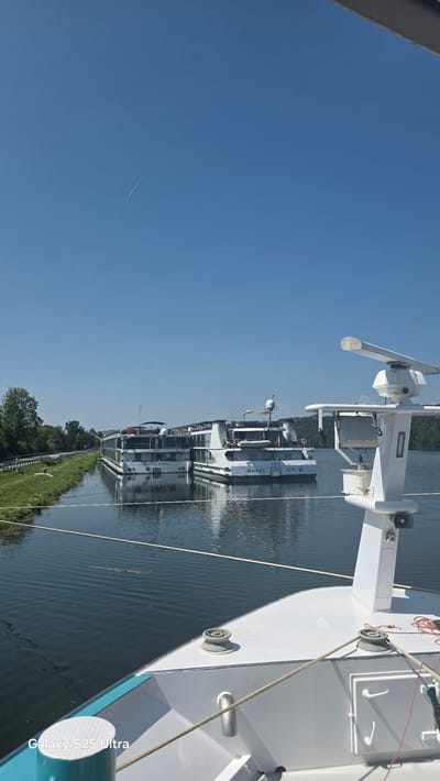 The image shows a clear blue sky with a faint contrail from an airplane. Below the sky are two large white river cruise ships docked side-by-side on a calm river. The ship on the right has "JOY BASEL CH" printed on its hull, and the ship on the left has "AMADEUS THENE" printed on its hull. The ships are moored in what appears to be a river port in Passau, Germany, as indicated by the presence of the Swiss flag on the "JOY" ship and its name referencing Basel. The riverbanks are lined with green grass and trees. In the foreground, the bow of another white boat is visible, equipped with nautical equipment like a radar antenna and cleats. The water is reflective, mirroring the sky and the ships.