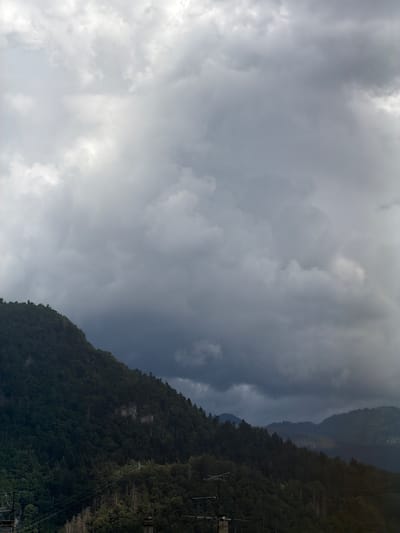 This image depicts a dramatic, cloudy sky over a lush, forested mountain range. The sky is filled with a mix of dark, ominous storm clouds and lighter, brighter patches, suggesting either the approach or departure of a storm. The mountains are densely covered in trees, their dark green foliage creating a stark contrast with the gray sky.
In the foreground, towards the right side of the image, several man-made structures are visible. These appear to be parts of a power transmission line or possibly a ski lift infrastructure, with poles and wires stretching up the mountainside. The presence of these structures, combined with the mountainous terrain, suggests a location that blends natural beauty with human development, potentially a recreational area or a mountain town.
The time of day is difficult to pinpoint precisely due to the overcast conditions, but the lighting suggests it is likely daytime, possibly late afternoon as the light seems to be fading slightly through the clouds. The overall mood is one of awe and perhaps a touch of melancholy, evoked by the powerful presence of nature and the dramatic weather. There are no people or animals visible in the image, and no text is present. The location context provided, Cluses, France, a town known for its Alpine setting and industry, aligns with the visual elements of mountains, forests, and infrastructure.