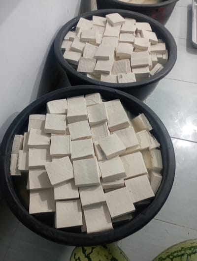 This image captures a close-up view of two large, black, circular tubs, both brimming with numerous white, square-cut blocks, which appear to be tofu. The tofu blocks are submerged in a clear liquid, likely water or brine, for preservation. The tubs are positioned on a light-colored, tiled floor, which shows reflections from an overhead light source.
In the background, on the right side, parts of two green watermelons with darker mottled patterns are visible, suggesting a market, kitchen, or food storage environment. A metallic tray or container is also partially visible in the upper left corner of the frame.
The tofu blocks vary slightly in their precise cubic shape, indicating they have been manually cut. They are densely packed within the tubs, forming a textured surface. The overall scene suggests a practical, functional setting related to food preparation or retail, specific to Dayahtuha, Indonesia. There are no people visible, nor any discernible text or prominent emotions. The lighting is artificial, illuminating the scene brightly.