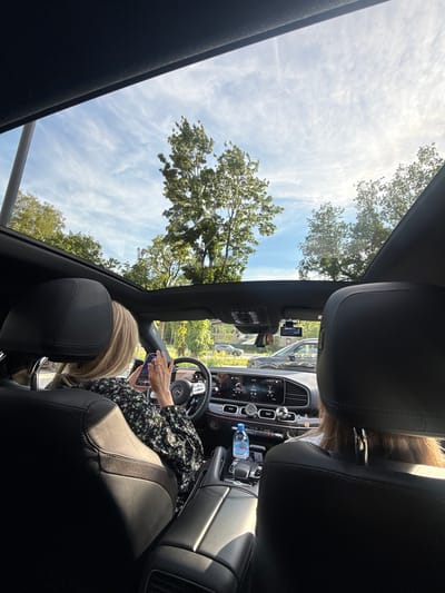 The image shows the interior of a car, viewed from the back seat, looking towards the front. Through the panoramic sunroof, a bright blue sky with wispy white clouds and lush green trees can be seen. The sun is high in the sky, suggesting it is daytime.

Inside the car, a person's hands are visible holding a smartphone. They are wearing a floral patterned dress. The dashboard of the car is visible, with its complex array of buttons and screens, indicating a modern vehicle. A bottle of water is placed in a cup holder. The headrests of the front seats are prominent in the foreground. The overall atmosphere appears relaxed and casual, possibly during a scenic drive. The location is identified as Zhukovka, Russia. No discernible text is visible in the image.