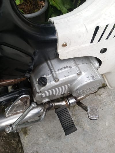 The image presents a close-up, angled view of the lower-midsection of an older model Suzuki motorcycle or scooter, located in Salai Har Htan, Myanmar. The vehicle is stationary on a grey concrete surface. Essential details include a silver-colored engine casing, heavily worn with visible scratches and pitting, featuring a black circular cap on its left side and the faint embossed "SUZUKI" logo. A black, ribbed rubber footpeg extends from the engine assembly, alongside a metallic brake or gear shifter pedal with a textured surface, both showing significant wear and some corrosion. Above the engine, a segment of the motorcycle's black frame or bodywork is visible, connecting to a faded white plastic side panel equipped with vertical ventilation slits and secured by a single, rusted screw. In the blurred background, green foliage and a dark potted plant are discernible, indicating an outdoor environment. No individuals are present, and no actions or interactions are depicted.