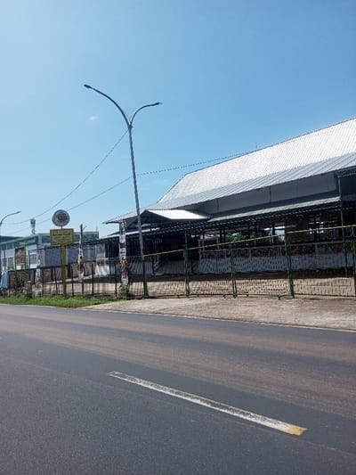 The scene depicts an outdoor urban environment under a clear, bright blue sky with minimal cloud cover. A two-lane asphalt road, partially covered with light-colored dust or debris, extends from the lower left towards the middle right. A solid white line with a shorter solid yellow segment marks a lane boundary on the road.
Adjacent to the road on the right is a narrow grassy verge, followed by a concrete sidewalk or paved area. A large building complex occupies the background. This complex features a wide, multi-gabled roof constructed from reflective metallic panels, indicative of a market or large public structure. Below the roof, an open-sided structure with visible support beams and pillars extends the length of the building.
A dark green metal fence, featuring a lower section with a brick-like pattern, runs along the front of the building complex, separating it from the sidewalk. Multiple vertical banners with faded graphics and text are affixed to posts along this fence. A prominent yellow rectangular sign, displaying an emblem and black text in a foreign script (including phrases like "SELAMAT DATANG DI PASAR MODERN BARADATU"), is visible behind the fence on the left. A large, circular, yellow and orange sign is partially visible to its left. Further left, a portion of a green and white building is present.
Two modern streetlights with double light fixtures are visible, one prominently in the upper middle and another partially on the far left. Dark electrical wires or cables stretch horizontally across the scene. A single individual is faintly discernible walking near the green and white building on the far left edge of the image.
The location is Beteng_Pangkhar_Somthrang, Bhutan.