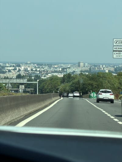 The image captures a view from a vehicle traveling on a highway, with the city of Châtenay-Malabry, France, visible in the background. A white Renault Megane is in the foreground, driving on the right side of the road. Other vehicles, including a white Peugeot 208 and a black Renault Clio, are also on the highway. Several traffic signs are visible, including a sign indicating "N 28 VERR CHÂTI -CENTI". The highway is bordered by a concrete barrier on the left and trees on the right. The sky is clear and blue.