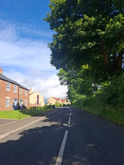 The image shows a road in Middleton Tyas, United Kingdom, with houses and trees lining the street. The road is asphalt with a white line down the center. On the left side of the road, there are modern brick houses with manicured lawns. A metal fence separates the properties from the sidewalk. On the right side of the road, there is a dense line of trees and greenery. The sky is blue with some wispy clouds, and the sun is casting shadows on the road. The scene appears to be a quiet residential area.