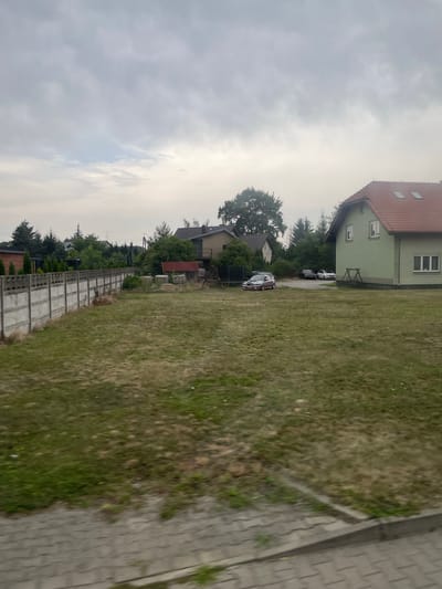 The image shows a residential area in Tyniec Mały, Poland, with a focus on a grassy yard bordered by a concrete fence. A light green house with a red tiled roof stands on the right side of the image. In front of the house, a small wooden swing set is visible. Several cars are parked on the property, including a silver hatchback and a dark sedan. Trees and smaller houses are present in the background, suggesting a suburban or semi-rural setting. The sky is overcast, with light gray clouds dominating the upper portion of the image. The foreground features a paved sidewalk and a lawn that appears to be dry and somewhat unkempt in places.