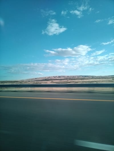 The image shows a view from a moving vehicle looking towards a landscape. The foreground features a dark asphalt road with a single yellow line running horizontally across it, indicating a lane divider. The road appears to be a highway. Beyond the road, a metal guardrail is visible, blurred due to motion. The middle ground shows a dry, arid landscape with rolling hills and sparse vegetation. The hills are layered with different shades of brown and tan, suggesting geological formations. In the distance, a faint horizon line separates the land from the sky. The sky is a clear, bright blue, with a few scattered white clouds. The overall impression is one of a roadside view in a somewhat desolate, natural environment. The location of Spassk, Kazakhstan is not discernible from the image itself.