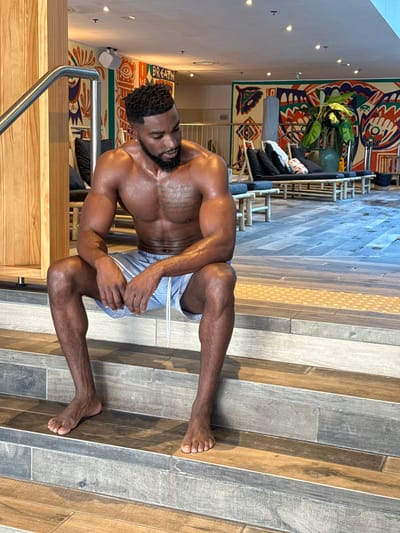An eye-level indoor shot depicts a muscular man, shirtless and wearing blue and white striped swim shorts, kneeling or crouching on a floor tiled with various shades of wood-look planks. His skin is visibly wet, suggesting he has recently been in water. He has a short beard, dark, curly hair, and tattoos on his chest. He looks directly at the camera with a neutral expression.
The setting is a modern leisure or spa facility, possibly in Levallois-Perret, France. In the background, a large, colorful tapestry with abstract patterns in orange, green, blue, and white adorns a white wall. A large potted plant with broad green leaves sits in front of the tapestry. Below this, lounge furniture with black cushions is arranged. Small recessed lights are visible on the ceiling. A metal handrail and light wooden structures are partially visible in the foreground on the left. The word "BREATH" is vertically inscribed on the tapestry. The lighting is bright and even, indicating a well-lit indoor space during the daytime.