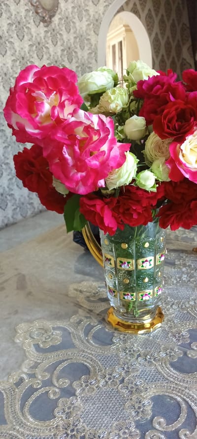 A vase filled with a bouquet of red and pink roses with white accents sits on a table covered with a lace tablecloth. The vase is made of clear glass with a gold base and intricate gold and floral patterns along its sides. In the background, a patterned wallpaper is visible. The scene appears to be an indoor setting in Tbilisskaya, Russia.