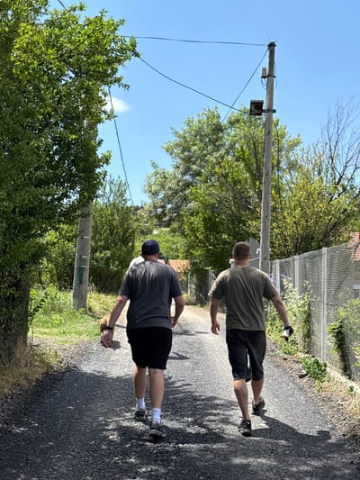 This image depicts two men walking down a sunny, tree-lined path in Grocka Urban Municipality, Serbia. Both men are viewed from behind, moving away from the camera.
The man in the foreground is wearing a dark grey t-shirt, dark shorts, white socks, and dark athletic shoes. He has a dark baseball cap on his head and holds a small, light-colored object in his right hand.
Further ahead, the second man wears an olive green t-shirt, dark shorts, and dark athletic shoes with light soles. He has short, light brown hair and appears to be wearing a light-colored glove on his left hand, holding a small white object, possibly a can or bottle.
The path is made of dark gravel or asphalt, illuminated by bright sunlight which casts strong, distinct shadows to the left of the walkers, indicating midday or early afternoon. The sky overhead is clear and blue. Green trees and dense bushes line both sides of the path, with a chain-link fence visible on the right. Concrete utility poles with wires are spaced along the left side of the path. The scene conveys a warm, sunny day in a natural, semi-rural setting. There is no visible text in the image.
