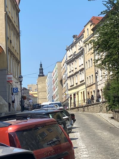 The image depicts a cobblestone street in Świdnica, Poland, on a clear day. The street is lined with buildings on both sides, characteristic of an older European town. On the left, a building with a light yellow facade features several windows and a red sign reading "Ergo Hestia". A blue parking sign is also visible. Several people are present: one individual stands near the "Ergo Hestia" sign, another walks down the street carrying a bag, and a third person is pushing a cart up the incline. On the right side, a multi-story building with a light yellow facade has numerous windows and a red-tiled roof. A large tree with green foliage partially obscures the building. In the foreground, several cars are parked along the street, including a red Dacia. In the distance, a dark-colored spire or tower is visible above the rooftops. The street slopes upwards, indicating it's an inclined road.