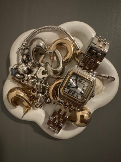 A white, irregularly shaped ceramic dish, approximately 20-25 cm at its widest point, is positioned on a plain, dark grey surface in an indoor setting in Wanneperveen, Netherlands. The dish contains a collection of jewelry and a wristwatch.

**Objects within the dish:**
*   A two-tone metal wristwatch with a square face, Roman numerals (IIII, V, VII, X, XI, XII visible), and a link bracelet is centrally placed, displaying approximately 10:10. The watch appears to be a Cartier Panthère model.
*   Multiple pieces of silver-colored and gold-colored jewelry are intermingled within the dish.
*   Silver-colored items include:
    *   A Pandora-style snake chain bracelet with various charms, including a charm resembling a Mickey Mouse head with blue enamel, and other patterned charms.
    *   A flat heart-shaped charm with the word "Niece" inscribed, featuring a small heart cut-out.
    *   Two pairs of silver-colored hoop earrings, one larger and one smaller.
    *   Several other small charms and a chain.
*   Gold-colored items include:
    *   Two pairs of gold-colored hoop earrings, one larger and one smaller, similar in style to the silver ones.
    *   A fine gold-colored chain.
    *   A gold-colored stud earring.
    *   A smooth, spherical gold-colored object, possibly a bead or another earring.
*   A small, light-colored seashell is partially visible beneath the watch on the right side.

No people are present in the scene, and no specific actions or interactions are depicted beyond the arrangement of the objects. The lighting is even, suggesting an interior photograph.