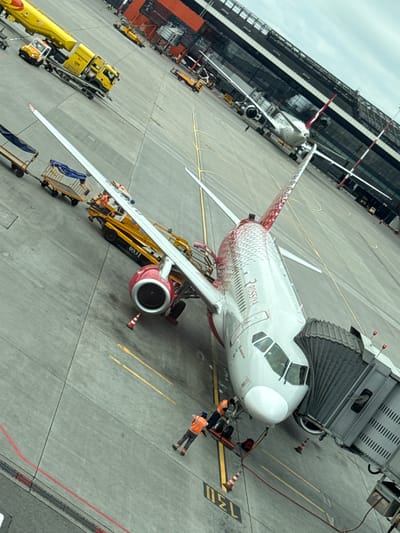 The image shows an airplane parked at an airport gate. The airplane is white with red accents and has "ROSSIYA" written on its side. A jet bridge is connected to the aircraft. Several airport vehicles, including yellow service trucks and baggage carts, are visible on the tarmac. Two individuals wearing orange safety vests are working near the front of the plane, one appearing to be operating equipment. The airport terminal building, a modern structure with large glass windows, is visible in the background. The scene is set on a paved tarmac, with yellow and red lines marking the parking area. The sky is overcast. The image is captured from a high-angle perspective.