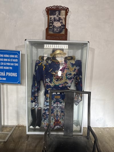 The image shows a display case mounted on a wall, containing historical artifacts. Inside the case, there is a traditional royal costume in dark blue with intricate gold and red patterns. Adjacent to the clothing are a pair of black shoes and a gold headpiece or crown. Mounted on the wall next to the display case is a framed photograph. Below the case, a blue informational sign with white text is visible. The Vietnamese text on the sign identifies the objects as being related to the Khải Định era and mentions Sà Phìn Commune, a location in Vietnam. The scene appears to be a historical exhibit in a museum or heritage site. The wall has a textured, painted surface. A portion of what looks like a metal structure is on the right side of the display.