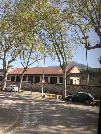 The image shows a street in Chambéry, France, lined with trees and buildings. The street curves slightly to the right. There are cars parked on the side of the road. The buildings are made of stone, some with red-tiled roofs. In the background, a mountain is visible. The image is taken from inside a vehicle, likely a bus or car. The time of day appears to be midday, with clear blue skies. There are no people visible in the image.
