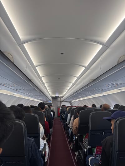 Inside an airplane cabin, passengers sit in rows of dark seats along a red-carpeted aisle. The plane's interior is predominantly white and gray, with curved walls illuminated by embedded lights. Passengers are generally facing forward, with their heads visible over the seatbacks. The perspective indicates a view from within the cabin, looking towards the front of the plane.