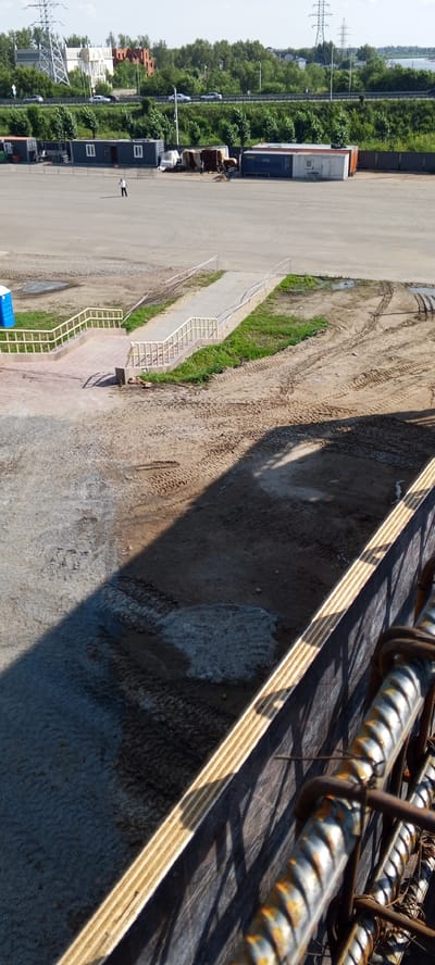 In Заречный ауылдық округі, Kazakhstan, on a bright, sunny day, a person is visible standing in an open, unpaved area. The ground is a mix of dirt and gravel, with tire tracks indicating vehicular activity. In the background, several modular buildings or shipping containers are arranged. One building has a blue portable toilet situated nearby. A paved walkway with yellow railings leads up to a higher level. The scene is dominated by an outdoor setting with a shadow cast across the ground, suggesting it is midday. The horizon shows a line of trees and utility poles. In the foreground, a construction element with rebar and wooden planks is partially visible, indicating ongoing work or an unfinished structure.