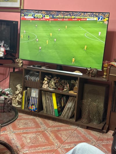 The image shows a television displaying a soccer game, with the score indicating "JANL 1" and "CHI 6" at the 50:26 mark of the first half. The TV is located in a room with a patterned tile floor and a brown wooden shelf filled with items, including decorative figures, CDs, and other objects. The overall environment suggests a residential setting. There are no explicit indications or recognizable landmarks to definitively place the location in San Cristóbal, Mexico; this location is inferred only based on details from the TV and OCR text. There are no people physically present in the frame.
