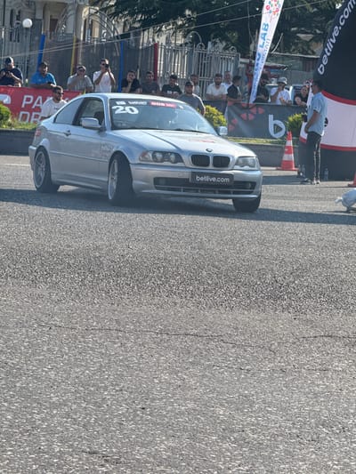 The image captures a street-level view of an outdoor event, likely a car race or show, in Poti, Georgia. A silver BMW sedan is prominently featured, positioned on an asphalt road. The car has several decals, including "betlive.com" on the front grille and the windshield, and a number "20" along with other graphics on the windshield and side.
A crowd of people lines the left side of the road, separated by a fence. They appear to be spectators, with some holding cameras and others observing the scene. The atmosphere seems lively, with people actively engaged. One person in the foreground is wearing a blue t-shirt, while others are dressed casually. There are also banners and flags visible behind the fence, adding to the event's festive ambiance.
The background shows some buildings and trees, suggesting an urban or semi-urban setting. The lighting indicates it is daytime, likely sunny, given the shadows and the bright asphalt. The overall scene depicts a community gathering centered around automotive activities, with commercial sponsorship evident from the visible branding.