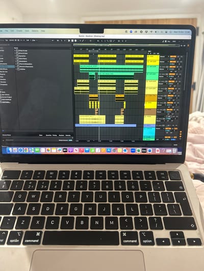 A silver-colored laptop, possibly a MacBook, is depicted from a slightly elevated frontal perspective, open and actively displaying software. The screen presents a Digital Audio Workstation (DAW) interface, specifically Ableton Live, with a project titled "Remix - Realism (Waking Up)". The interface shows multiple horizontal tracks with colored clips representing audio or MIDI data, labeled with elements such as "KICK", "HAT LOOP", "CLAP LOOP", "ZEN_TTB_12", "RISER", "VOCAL", and "Bpm128_Frees_Bass (1)". A mixer section with volume faders and other controls is visible on the right. The macOS menu bar at the top displays system icons, including the date and time "Wed 15 Oct 12:39", and a screen notch is present centrally. The macOS Dock is visible at the bottom of the screen with various application icons.
The laptop features a QWERTY keyboard with black keys and white legends, including a '£' symbol on the '3' key, indicating a United Kingdom layout. Visible dust and smudges are present on the keyboard keys and the silver casing surrounding the keyboard and trackpad.
The laptop rests on a light-colored, textured fabric surface, likely a bed or blanket, exhibiting visible folds. The background behind the laptop is softly blurred, revealing a light-toned wall or door frame. A dark cable with a metallic connector is partially visible, plugged into the right side of the laptop. No individuals are present in the image. The image provides no verifiable information regarding a specific location within Winslow, United Kingdom.