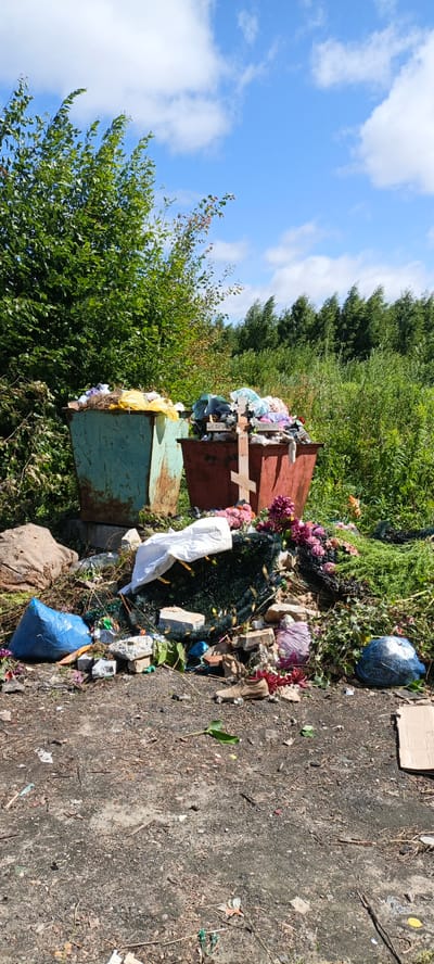 The image depicts an outdoor scene in Puchezh, Russia, characterized by two overflowing refuse containers situated amidst overgrown vegetation. The refuse containers, one teal and one reddish-brown, are filled with a mixture of discarded items, including textiles, plastic bags, and what appear to be artificial flowers. In front of the containers, a pile of debris has accumulated, comprising bricks, plastic sheeting, artificial greenery, and scattered floral arrangements, including a wooden cross. A single brown shoe rests on the ground amongst the trash. The surrounding environment consists of dense green foliage and trees under a blue sky with scattered clouds. The ground is a mixture of dirt and gravel, littered with smaller pieces of refuse.
