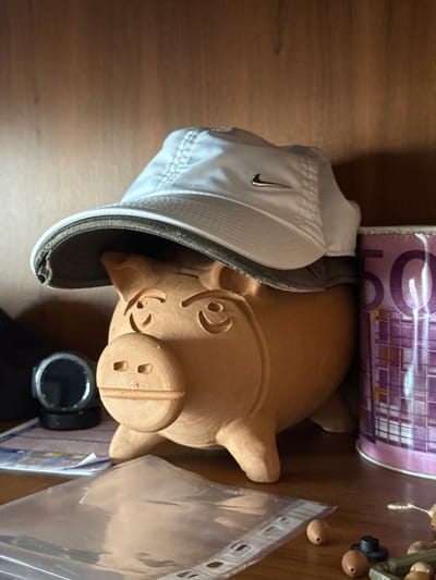 A still-life image is shown, oriented vertically, displaying a collection of objects on a wooden surface. The primary subjects are a small, unglazed terracotta pig figurine, a white baseball cap with a Nike logo, and a rolled-up bank note. The pig figurine is positioned on its side, with its snout facing towards the right edge of the frame. The baseball cap is placed over the pig, with its brim obscuring part of the figurine's back. The rolled bank note, which appears to be a 500 Euro note due to the visible purple color and numerals, rests behind the pig.
Other objects visible include a clear plastic sleeve with punched holes, and a few small, round, wooden beads. A black digital watch with a round face is also present, lying near the bottom right of the frame. The wooden surface suggests an indoor setting, possibly a desk or shelf. The lighting is somewhat dim, casting shadows and highlighting the textures of the objects. The image appears to be taken from a slightly elevated angle, looking down on the arrangement. There is no visible indication of time of day, weather, or specific emotions conveyed by the arrangement. The visible text on the bank note is primarily the numeral "500" and purple patterned designs. The Nike logo on the cap is also legible. The location context provided is Bova Marina, Italy, but there are no explicit visual cues within the image to confirm this.