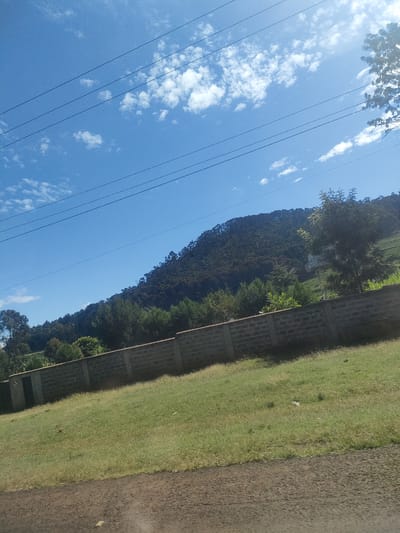 The image depicts a scene in Kibirichia ward, Kenya. A stone block wall runs horizontally across the midground of the image, with a grassy area extending from the wall towards the bottom of the frame. Beyond the wall, there are trees and a hill densely covered with trees. The sky is visible in the upper portion of the image, featuring a bright blue color with scattered white clouds. Power lines are also present against the sky. The photograph appears to be taken from a slightly angled perspective, potentially from a moving vehicle.