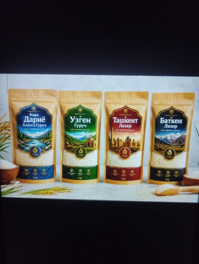 The image displays four bags of rice arranged horizontally. The bags are predominantly light brown with colorful labels featuring various landscapes and text in what appears to be Cyrillic script. Each bag is branded with "PREMIUM QUALITY" and has a distinct color scheme and design on its label.
From left to right:
1. A bag with a blue and gold label depicting a river flowing through a mountainous landscape. Text includes "Кора Дариё Аланга Гуруч 100% Таиийн Махсулот". At the bottom, there's a blue band with text and "1 кг".
2. A bag with a green and gold label showing a village scene in a mountainous region with traditional architecture. Text includes "Узген Гуруч 100% Таиийн Махсулот". At the bottom, there's a green band with text and "1 кг". The city of Özgön is mentioned in the text on this bag, suggesting a connection to the product's origin or branding.
3. A bag with a red and gold label featuring an image of grand Islamic architecture, likely in a city setting. Text includes "Ташкент Лазер 100% Таиийн Махсулот". At the bottom, there's a red band with text and "1 кг".
4. A bag with a dark blue and gold label showcasing a majestic mountain range under a clear sky. Text includes "Баткен Лазер 100% Таиийн Махсулот". At the bottom, there's a dark blue band with text and "1 кг".
To the left of the first bag, a portion of a wooden bowl filled with white rice and a few stalks of rice are visible. To the right of the last bag, a wooden scoop is placed on a pile of rice, and a glimpse of another wooden bowl is visible. The background is a textured, light-colored surface, possibly a table or countertop, with some soft lighting. The overall presentation is that of a product advertisement.