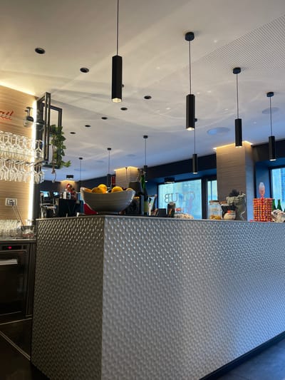 The image displays the interior of a modern bar, café, or lounge, oriented vertically but depicting a horizontal space. A prominent, long bar counter is on the right side of the frame, featuring a distinctive light gray, possibly metallic, hammered or dimpled texture on its front panel.
Above the bar and across the ceiling, numerous black hexagonal pendant lights hang at varying lengths, interspersed with circular recessed lights. Parts of the ceiling also show perforated acoustic panels.
Behind the bar, a variety of items are arranged for service. These include several bottles, a bowl filled with yellow citrus fruits (likely lemons or oranges), and other bar-related equipment. A black multi-tiered shelf below the ceiling further displays potted green plants and rows of inverted clear stemware hanging from racks. In the background, large windows allow natural light to enter the space, and a reflected sign displaying the word "CLUB" (backward) is visible.
No people are present in the scene, suggesting a quiet period in the establishment. The combination of natural light through the windows and the active interior lighting indicates it is daytime. The overall aesthetic is clean, contemporary, and functional, consistent with a modern hospitality venue. No specific emotions are discernible.