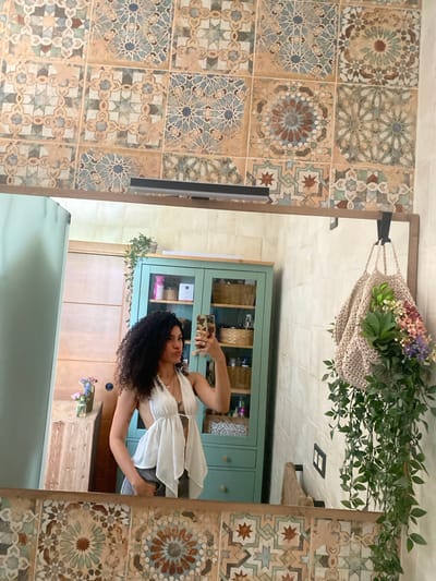 The image shows a woman taking a selfie in a bathroom. She is wearing a white top and grey pants. Her hair is dark and curly. The bathroom has patterned tiles on the walls, a green cabinet, and a wooden counter. There is also a mirror with a macrame bag filled with flowers and greenery hanging from it. The overall atmosphere is relaxed and casual, suggesting a daytime setting. The presence of tiles and a cabinet indicates an indoor domestic space, likely a bathroom or vanity area. The woman's relaxed pose and the selfie suggest she is comfortable and perhaps documenting her outfit or surroundings. There is no visible text in the image.