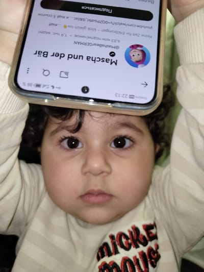 A close-up shot features a young child lying down, facing the camera with wide, curious eyes. A smartphone is held near the child's head, displaying a YouTube channel page. The channel "Mascha und der Bär" (@MashaBearGERMAN) has 6.83 million subscribers. The child wears a striped shirt with "MICKEY MOUSE" stitched on the sleeve. The phone's screen shows the video "Keine Zeit für Erklärungen - klick gleich los" and a "Подписаться" (Subscribe) button.