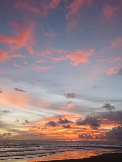 The image displays a sunset over a coastal area in Tibubiyu, Indonesia. The sky occupies approximately two-thirds of the frame, transitioning from a pale blue at the top through light grey, then exhibiting extensive pink and orange cloud formations across the mid-sky. Near the horizon, intense orange and red light from the setting sun dominates, with darker, more defined grey clouds interspersed within this vibrant band, particularly towards the right. The ocean occupies the middle ground, appearing dark blue-grey with several visible lines of white-crested waves approaching the shore. The immediate foreground consists of wet, dark sand on the beach, which reflects the warm orange and red colors of the sunset, creating a mirror-like effect. No individuals, animals, or man-made objects are visible within the frame.