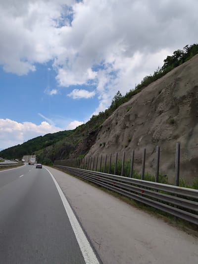 The image depicts a highway scene near Malo Buchino, Bulgaria. A multi-lane highway curves to the left, with cars visible in the distance. To the right of the highway, a steep, rocky hillside is reinforced with netting and vertical posts. A metal guardrail runs along the edge of the road. The sky is blue with scattered white clouds. The vegetation on the hillside is sparse, with some trees and bushes visible at the top.